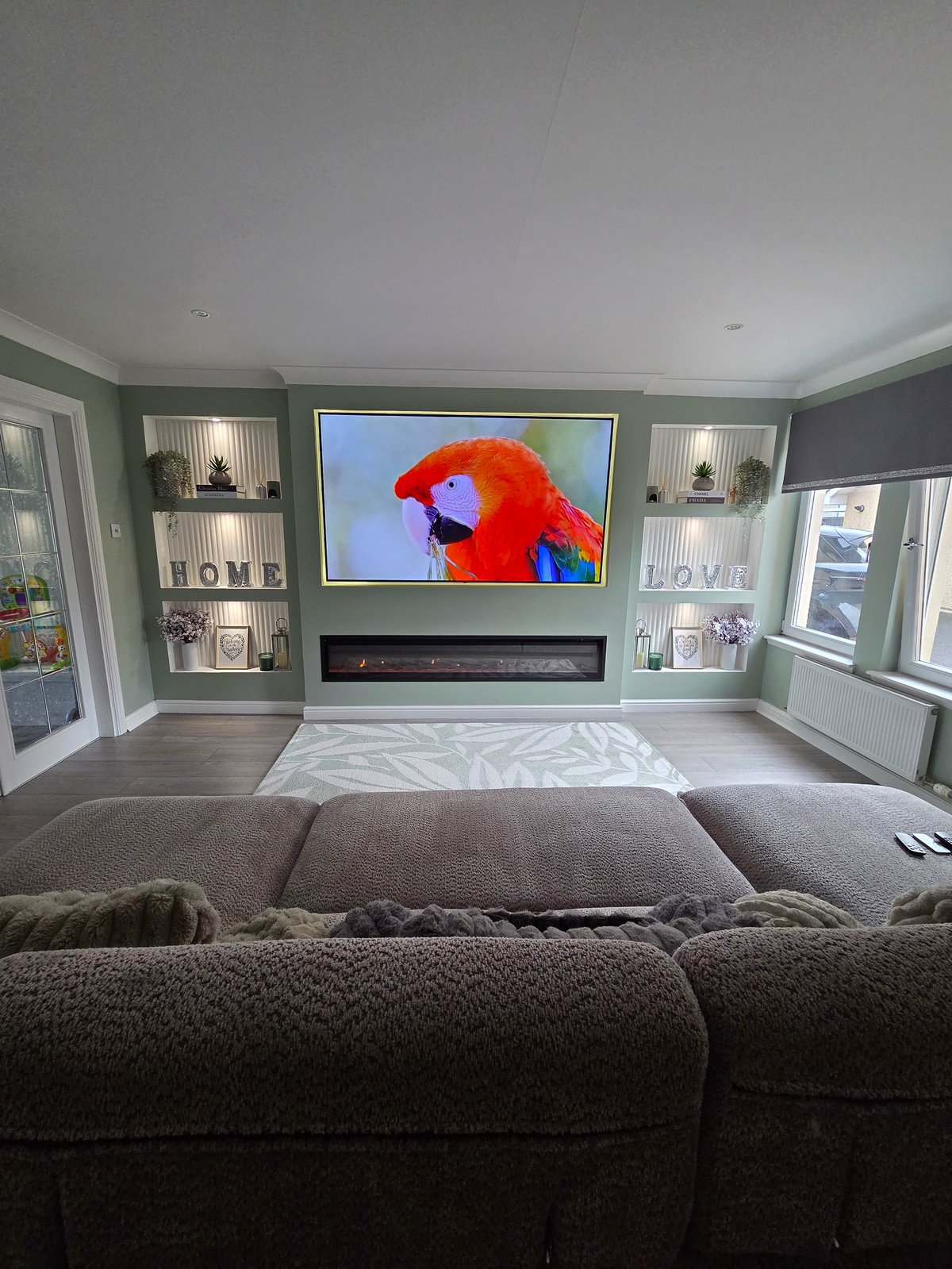 Modern living room with wall-mounted TV, sage green accent wall, built-in shelving with HOME decor, upholstered bench seating and warm ambient lighting