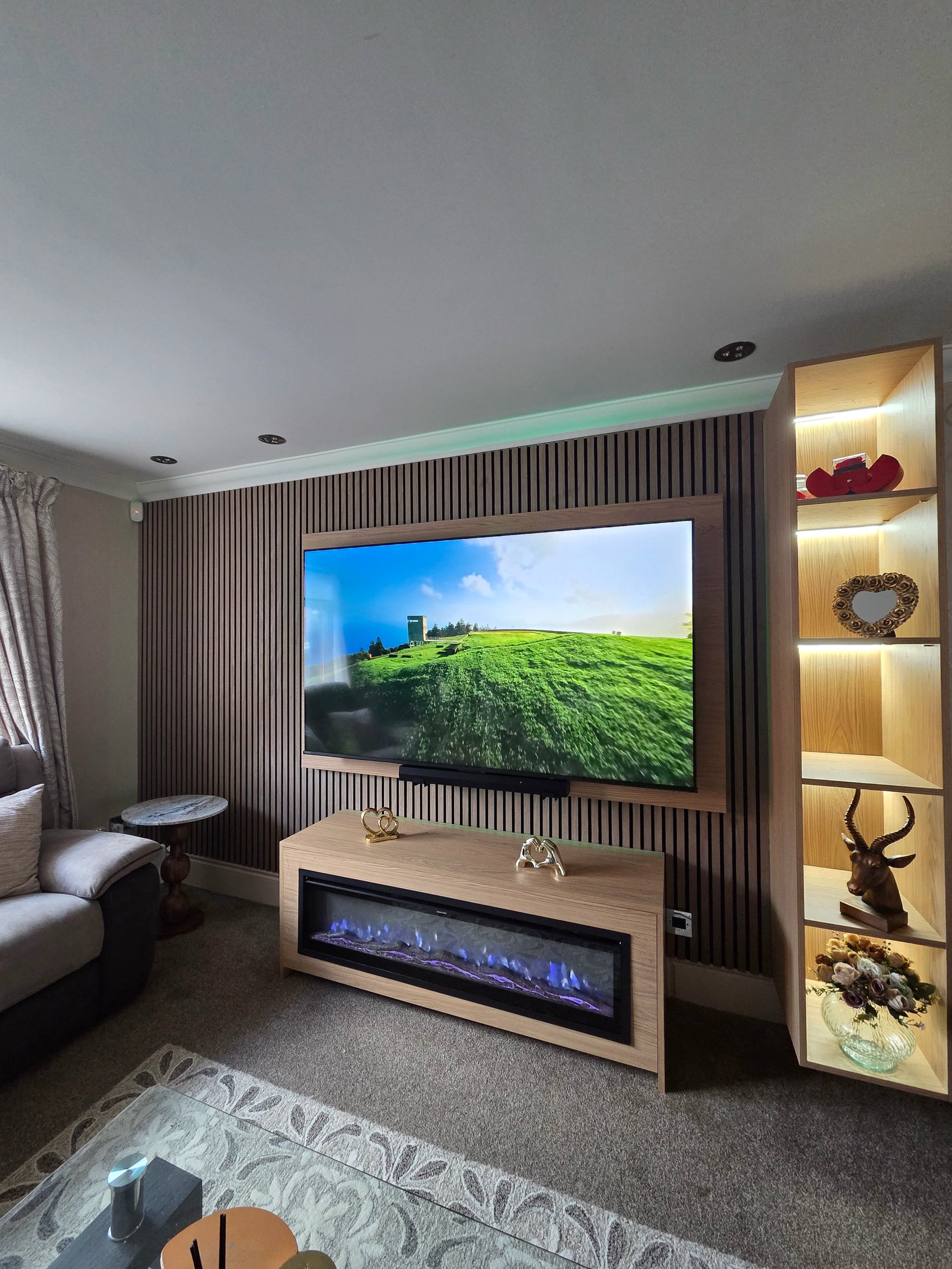 Modern home theater room with wall-mounted TV displaying a landscape scene, electric fireplace below, illuminated wall shelving with decorative items, and contemporary seating