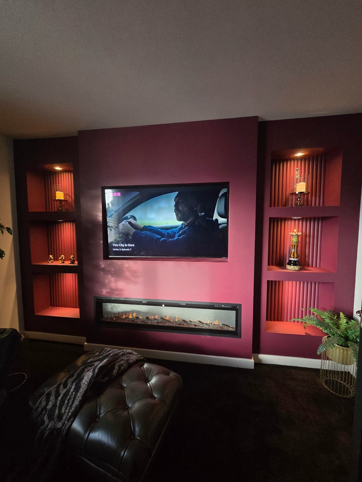 Modern living room with pink accent wall, mounted TV, glowing shelving, black leather seating, and warm ambient lighting