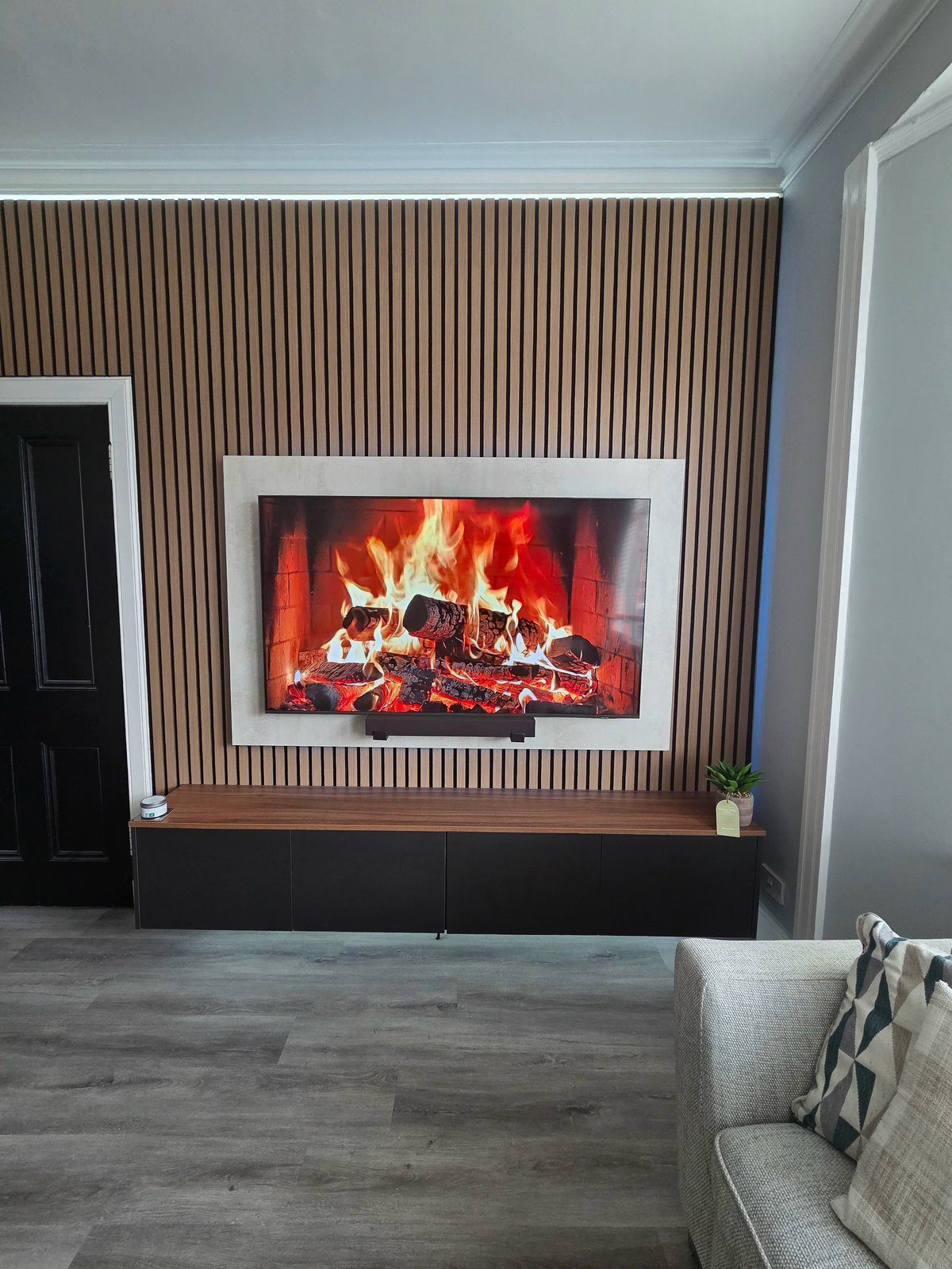 Modern living room with electric fireplace displaying flames, mounted on corrugated wood accent wall with white frame, dark base, and cream armchair in foreground